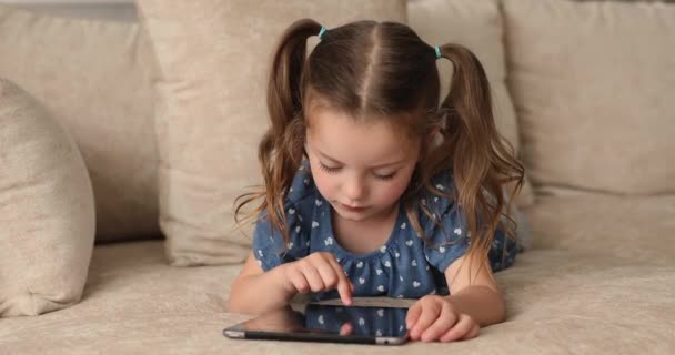 Маленька дівчинка лежить на дивані грати в відеоігри на планшеті — стокове відео