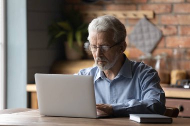 Eski erkek uzaktan kumandalı çalışan mutfak masasında bilgisayar kullanarak oturuyor.