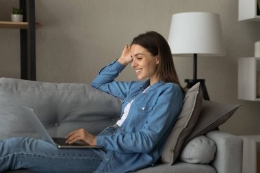 Yan gözle gülümseyen kadın dizüstü bilgisayar kullanıyor, koltukta oturuyor.