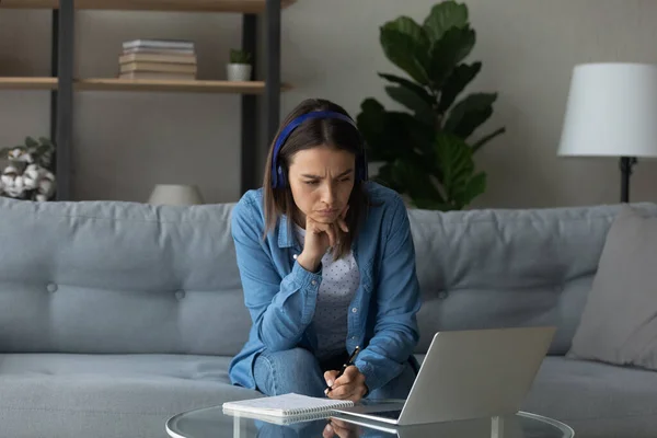 Focused woman wearing headphones looking at laptop screen, taking notes — Stock Photo, Image