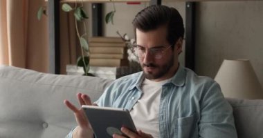 Gözlüklü adam kanepeye oturup tablet aleti tutuyor.