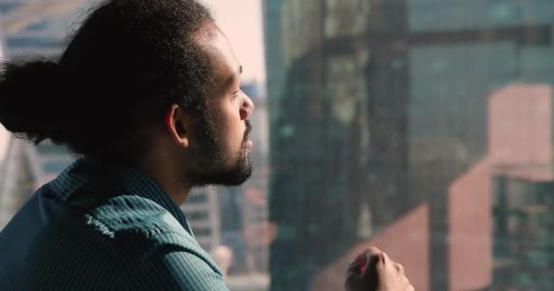 El hombre africano pensativo mira por la ventana a los rascacielos modernos de la ciudad — Vídeo de stock