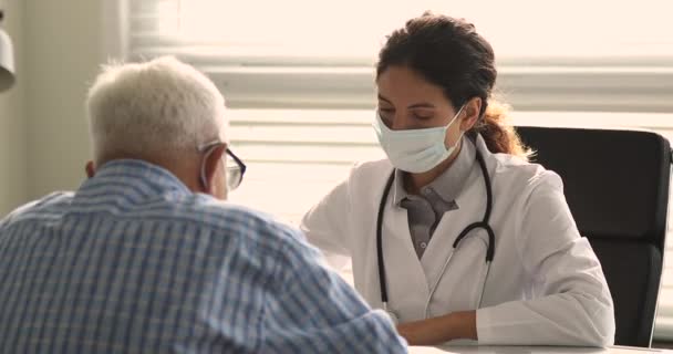 Leende vänlig läkare i ansiktsmask konsultera patienten vid kontrollmöte. — Stockvideo
