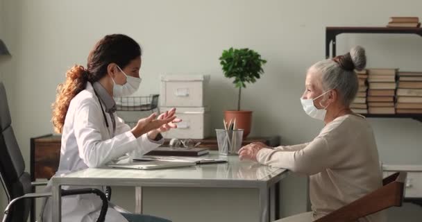 Uppmärksam medelålders äldre kvinna och ung läkare i ansiktsmasker diskuterar behandling. — Stockvideo