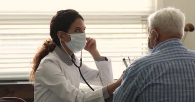 Yüz maskeli profesyonel kadın terapist eski bir hastanın akciğerlerini kontrol ediyor..