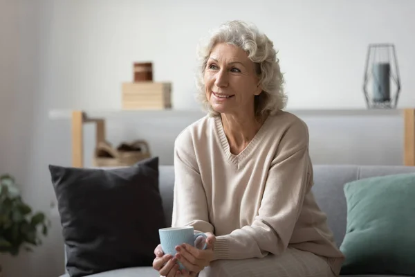 Smiling pensive mature woman holding cup of tea or coffee