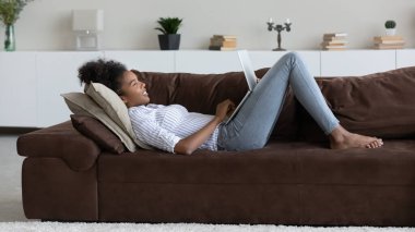 Genç melez kadın dizüstü bilgisayar kullanarak kanepede dinleniyor.
