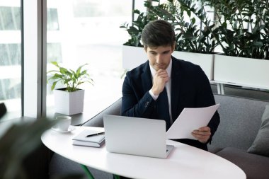 Düşünceli iş adamı elinde kağıt belgelerle dizüstü bilgisayarda çalışıyor.