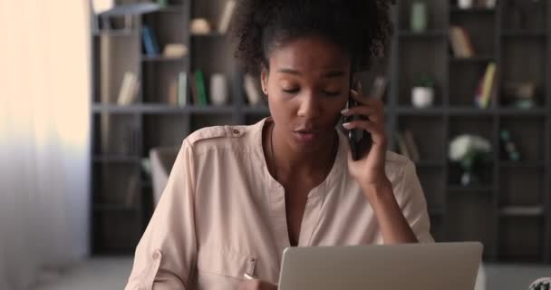 Koncentreret ung afrikaner kvinde taler på mobiltelefon, arbejder på computer. – Stock-video