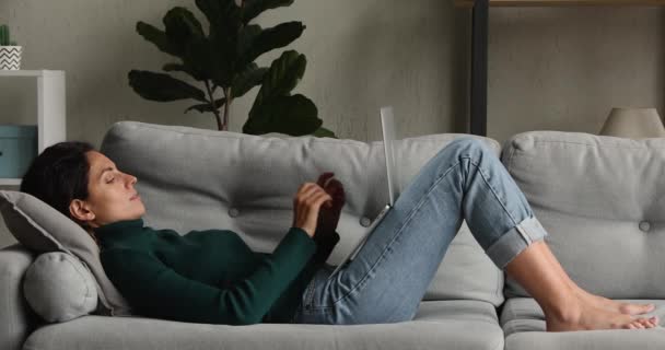 Jeune femme heureuse détendue tapant sur un ordinateur portable, allongée sur un canapé. — Video