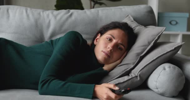 Δυστυχισμένη νεαρή γυναίκα ξαπλωμένη στον καναπέ, βαριέται βλέποντας τηλεόραση. — Αρχείο Βίντεο