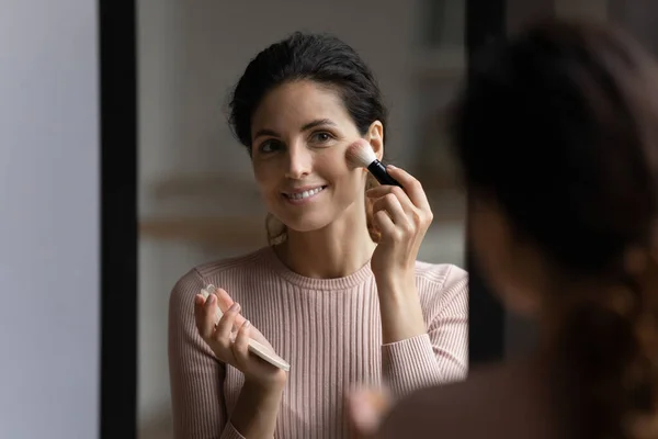 Pretty latina woman stand near mirror apply powder on face