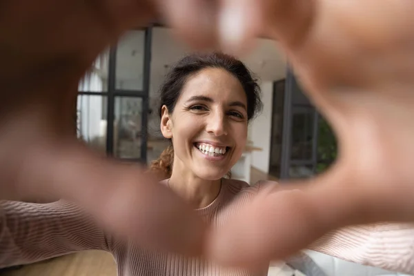 Hispanic kvinne ser på kamera gjennom hjertet av sammenføyde fingre. – stockfoto