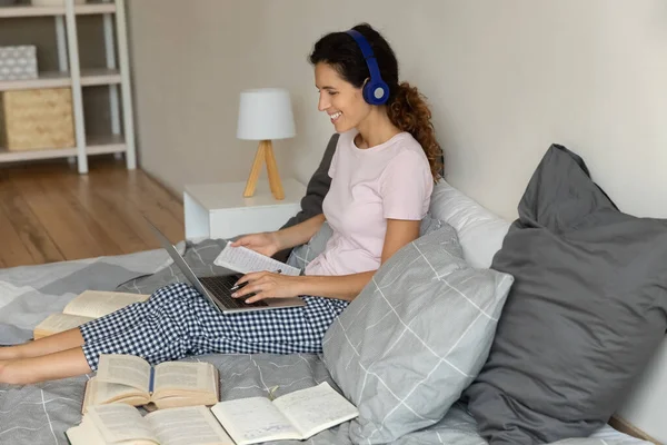 Young hispanic woman in earphones sit on bed by laptop