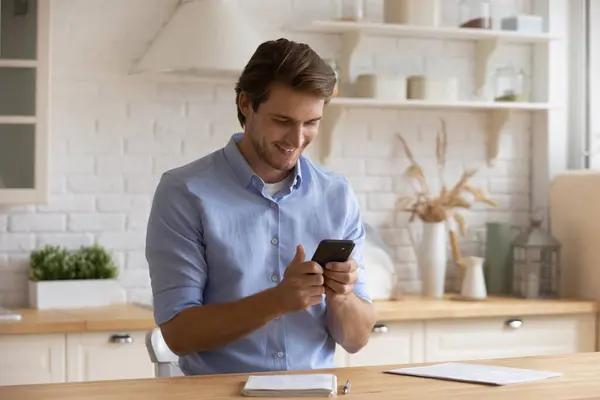 Smiling man holding smartphone, looking at screen, sitting in kitchen — Stock Photo, Image