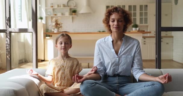 Mother her little daughter meditating seated cross-legged on couch — Stock Video