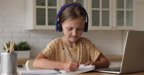 Schoolmeisje dragen hoofdtelefoon luisteren audio op laptop schrijft in copybook — Stockvideo