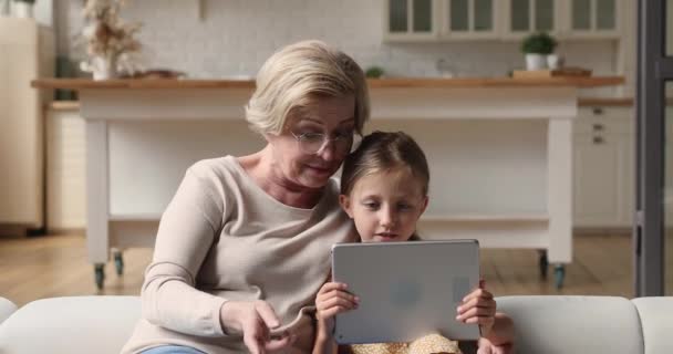 Старша бабуся і маленька онучка відпочивають на дивані з планшетом — стокове відео