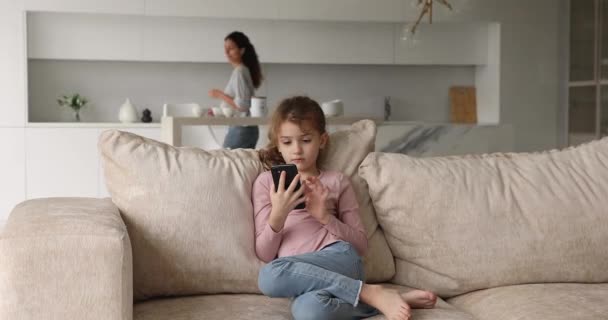 Дочка сидить зі смартфоном, поки її мати займається домашніми справами — стокове відео