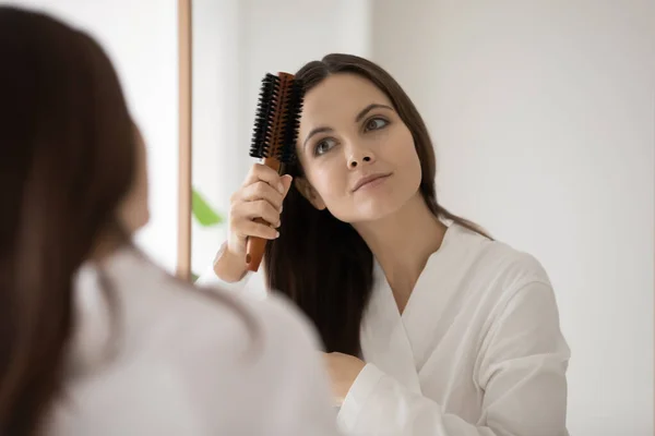 Menina em roupão escovando cabelo marrom liso no espelho — Fotografia de Stock
