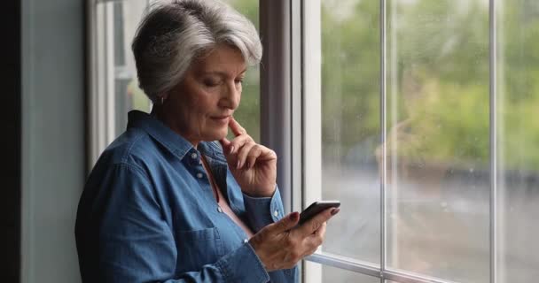 Sonriente anciana soñadora usando smartphone, de pie cerca de la ventana. — Vídeos de Stock
