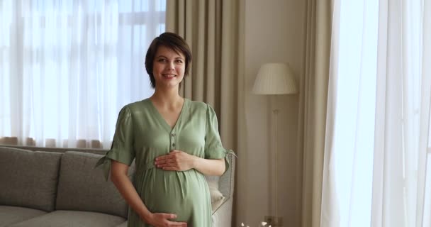 Frau, die drinnen steht, lächelt Bauch an und genießt Schwangerschaft — Stockvideo