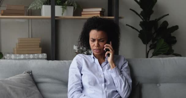 Frowning African woman sit on sofa swears on the phone — Stock Video