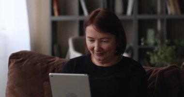 Yaşlı kadın kanepeye oturmuş elinde modern tablet elektronik cihazla.