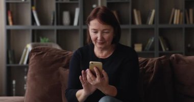 Yaşlı kadın elinde akıllı telefonuyla kanepede oturuyor.