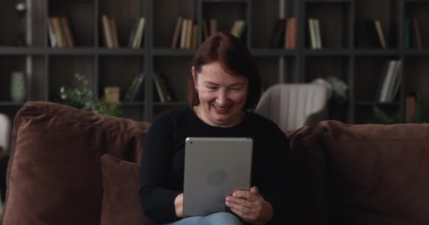 Ευτυχισμένη ηλικιωμένη γυναίκα κάθεται στον καναπέ χρησιμοποιώντας συσκευή tablet — Αρχείο Βίντεο