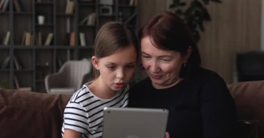 60 'ların büyükannesi ve küçük torunu elinde tabletle kanepede oturuyor.
