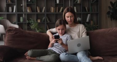Annesi dizüstü bilgisayar kızı kullanıyor. Akıllı telefonu kanepeye oturtuyor.