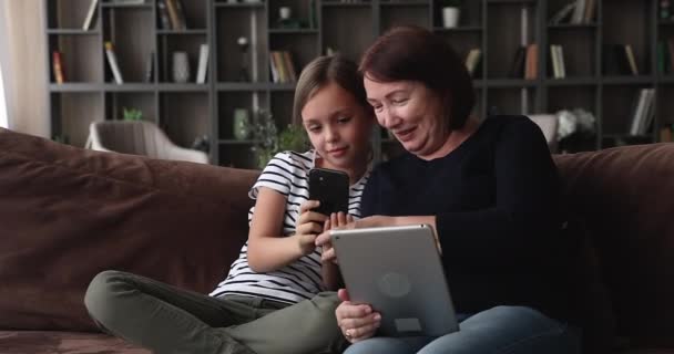 Abuela y nieta sosteniendo la tableta y el teléfono inteligente sentarse en el sofá — Vídeos de Stock