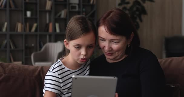 60s granny and little granddaughter sit on couch with tablet — Stock Video