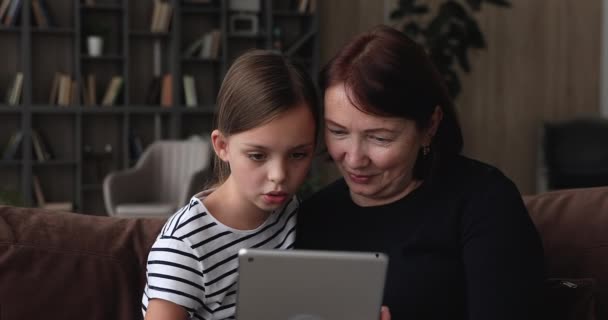 60 abuelita y nieta pequeña sentarse en el sofá con la tableta — Vídeos de Stock