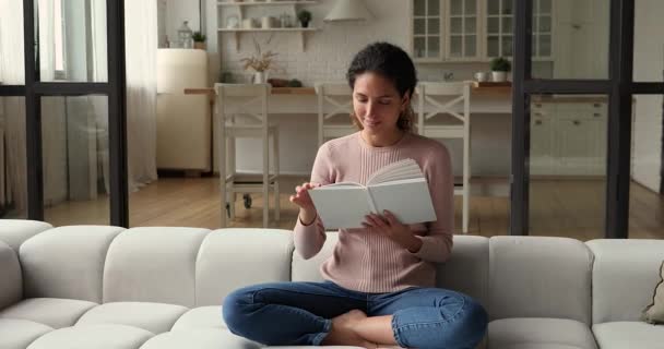 Young woman reading book seated cross-legged on couch indoor — Stock Video