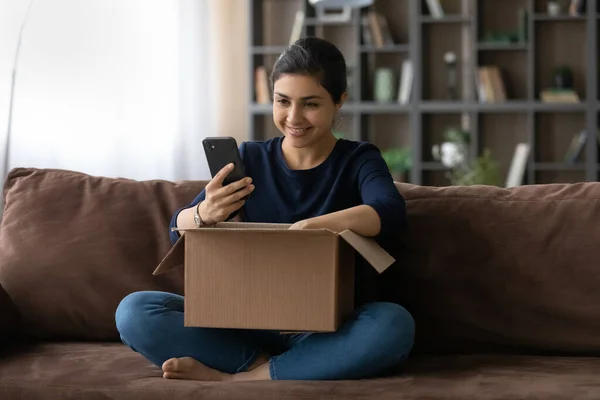 Usmívající se indická žena pomocí smartphonu, vybalování balíček doma — Stock fotografie