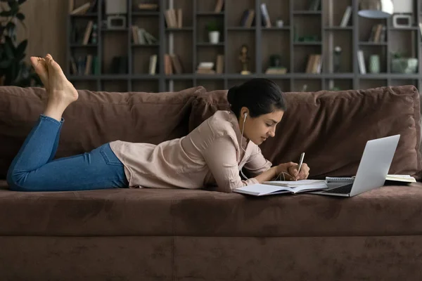 Side view Indian woman in earphones lying on couch, studying