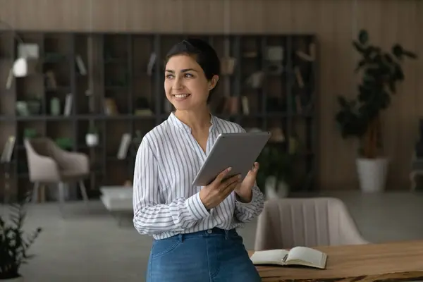 Dreamy smiling Indian businesswoman holding tablet, looking to aside
