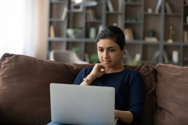 Confident Indian woman using laptop, sitting on couch, working online