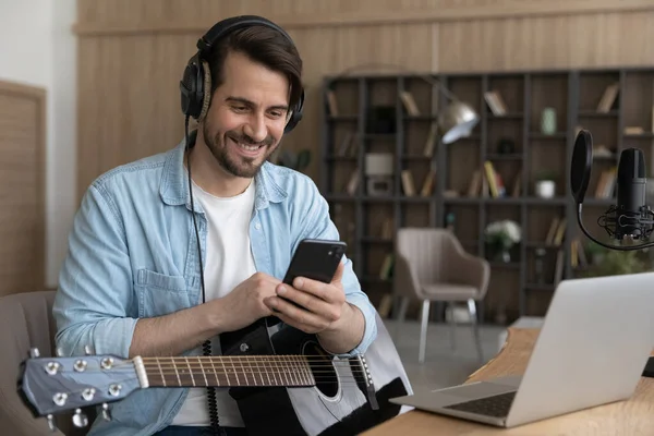Smiling man in headphones using smartphone, sitting with guitar — Stock Photo, Image