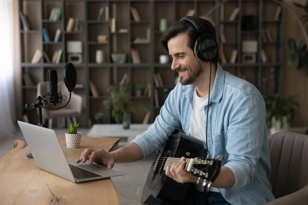 Smiling man in headphones using laptop, playing acoustic guitar — Stock Photo, Image