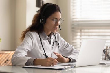 Kulaklıklı genç bayan doktor Webinar 'ın not almasını izliyor.