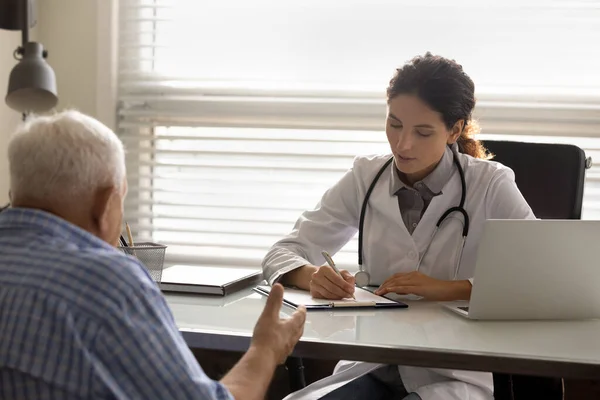 Médico atento femenino que escribe la historia clínica del paciente anciano del hombre — Foto de Stock