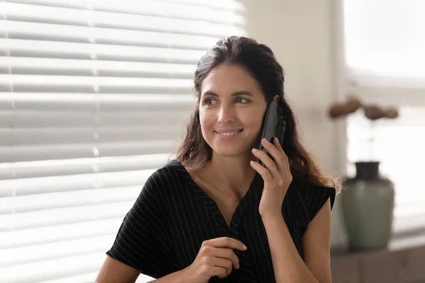 Щаслива молода іспаномовна жінка стоїть біля вікна, розмовляючи по телефону — стокове фото