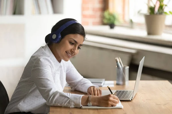 Sorrindo mulher indiana interessada em fones de ouvido tomar notas, usando laptop — Fotografia de Stock