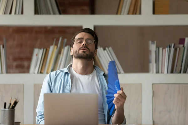 Exhausted overheated businessman in glasses waving paper fan in office