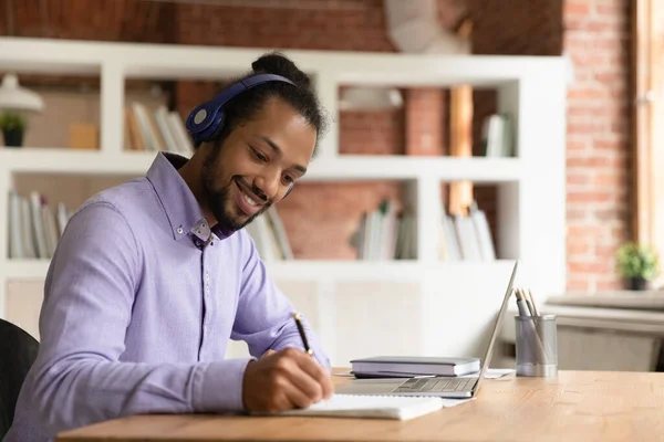 Smiling African American man in headphones taking notes, studying online — Stock Photo, Image