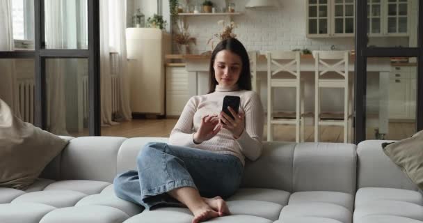 Hermosa mujer relajante en el acogedor sofá en casa con teléfono inteligente — Vídeos de Stock