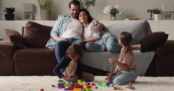 Gelukkige liefdevolle ouders kijken naar twee kinderen spelen op de vloer. — Stockvideo